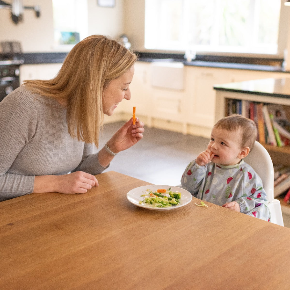Mother feeding baby, baby wearing long sleeve bib with fruits print sitting on a highchair