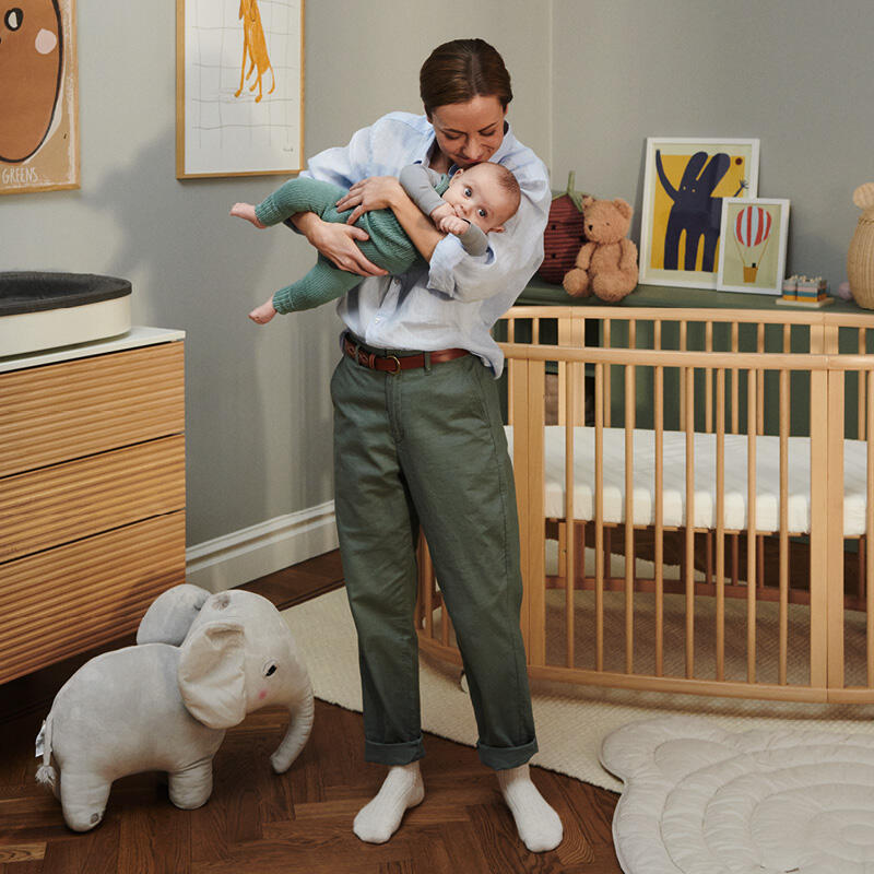 Woman holding a baby in a nursery with a crib, toys, and artwork.