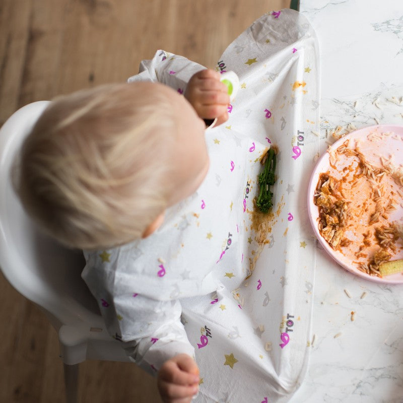 Boy wearing disposable bib sitting on highchair attached with dinning table eating food.