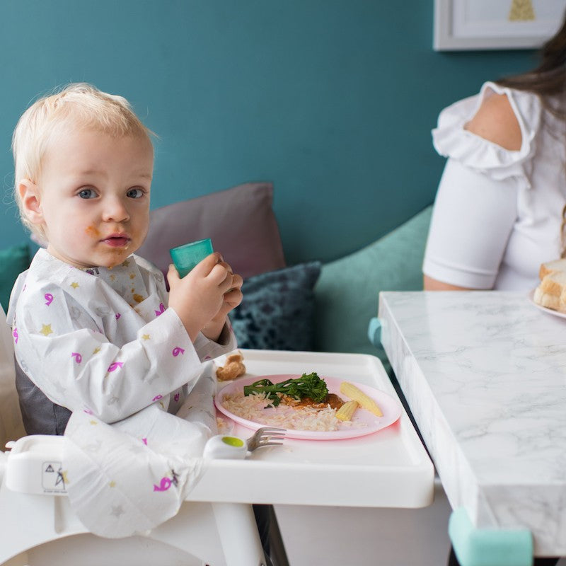 Boy wearing disposable bib sitting on highchair eating food.