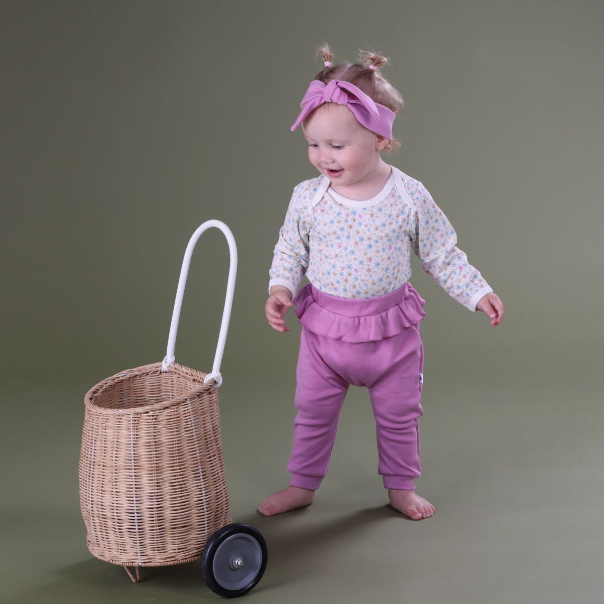 Baby girl standing with a basket wearing merino frill pants and head band pairing it with floral bodysuit.