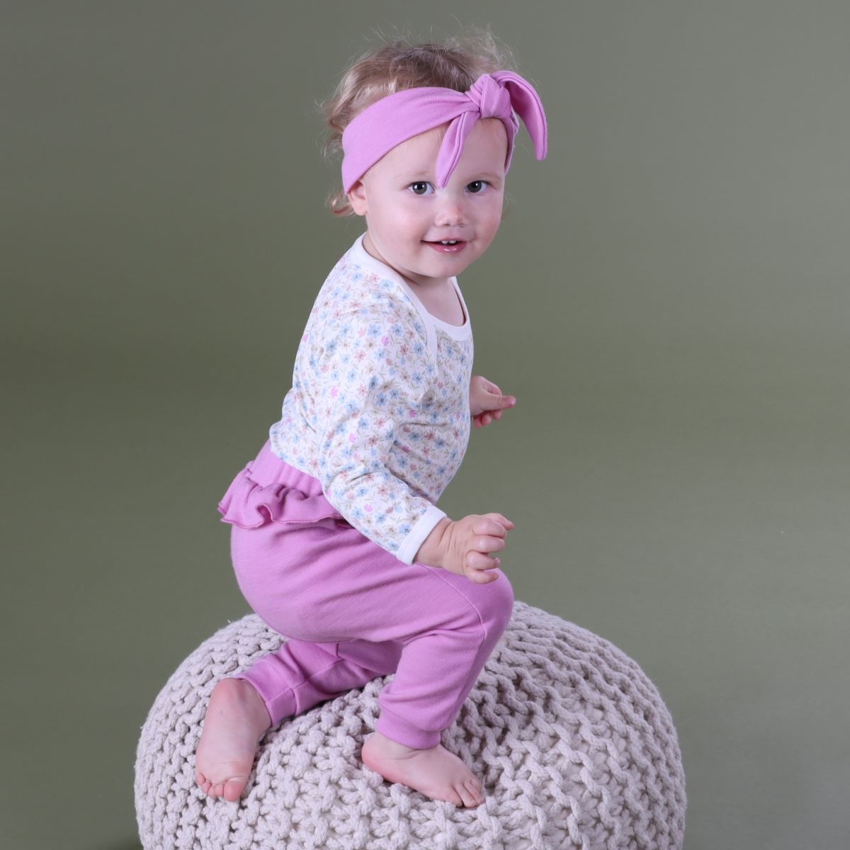Baby girl sitting in a pillow wearing merino frill pants and head band pairing it with floral bodysuit.