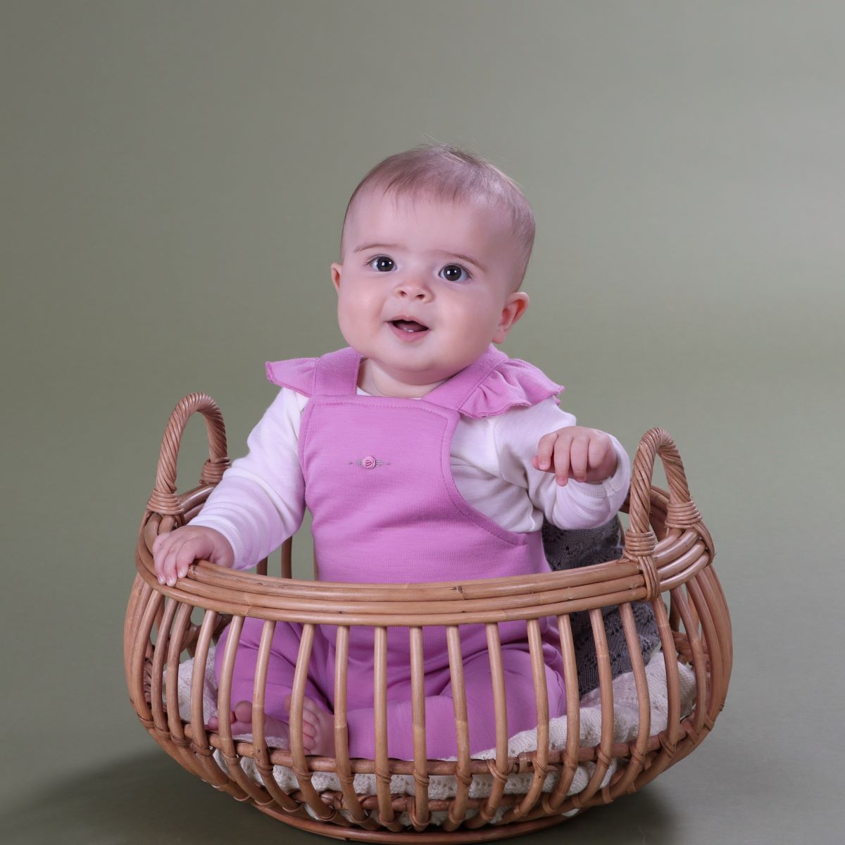 Baby wearing orchid merino frill overall and pairing it with merino long sleeve bodysuit sitting in a basket