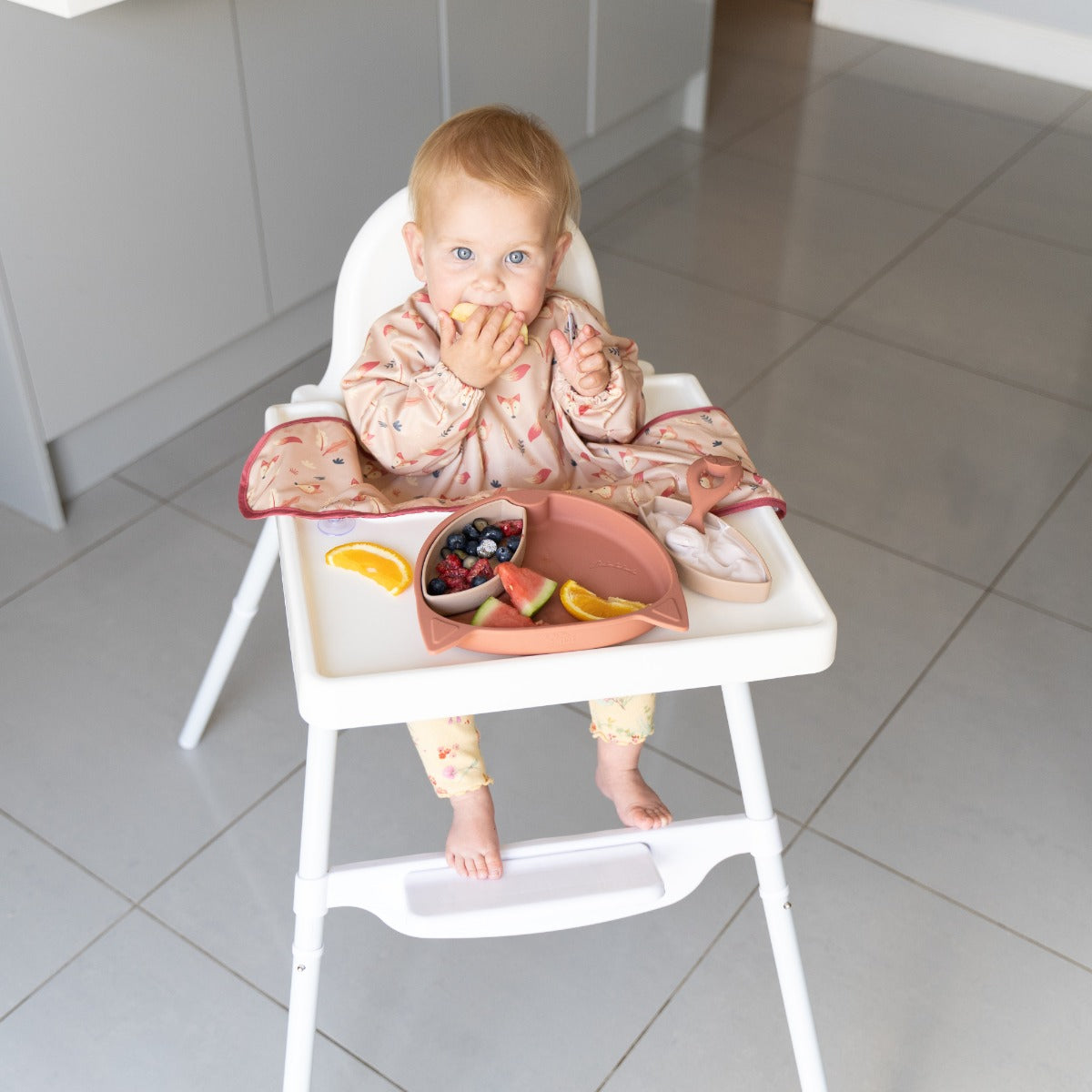 Baby wearing long sleeve bib with fox print eating as seated on a highchair