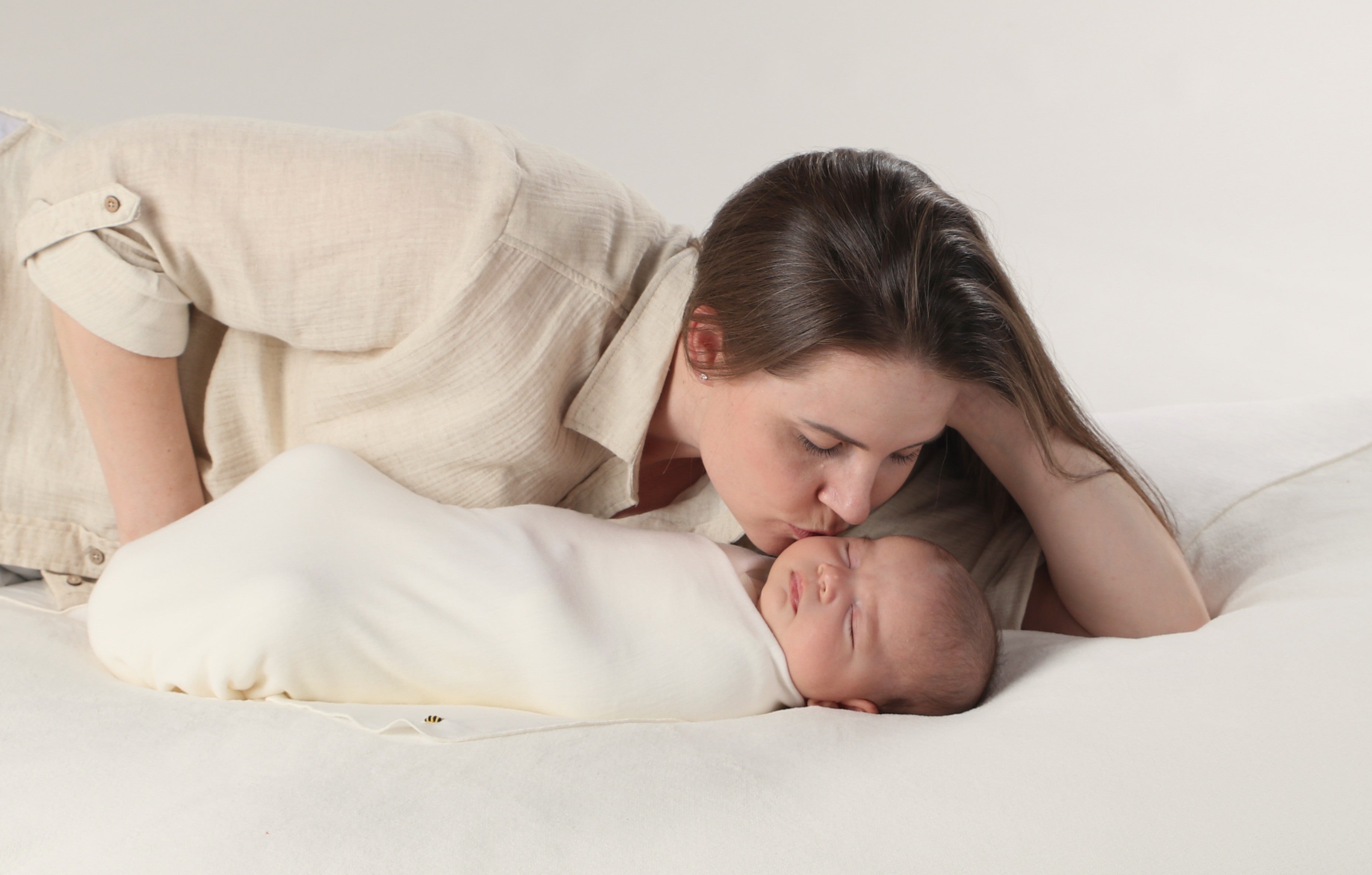 Woman lying on a bed with a baby wrapped in a white blanket, both looking peaceful.