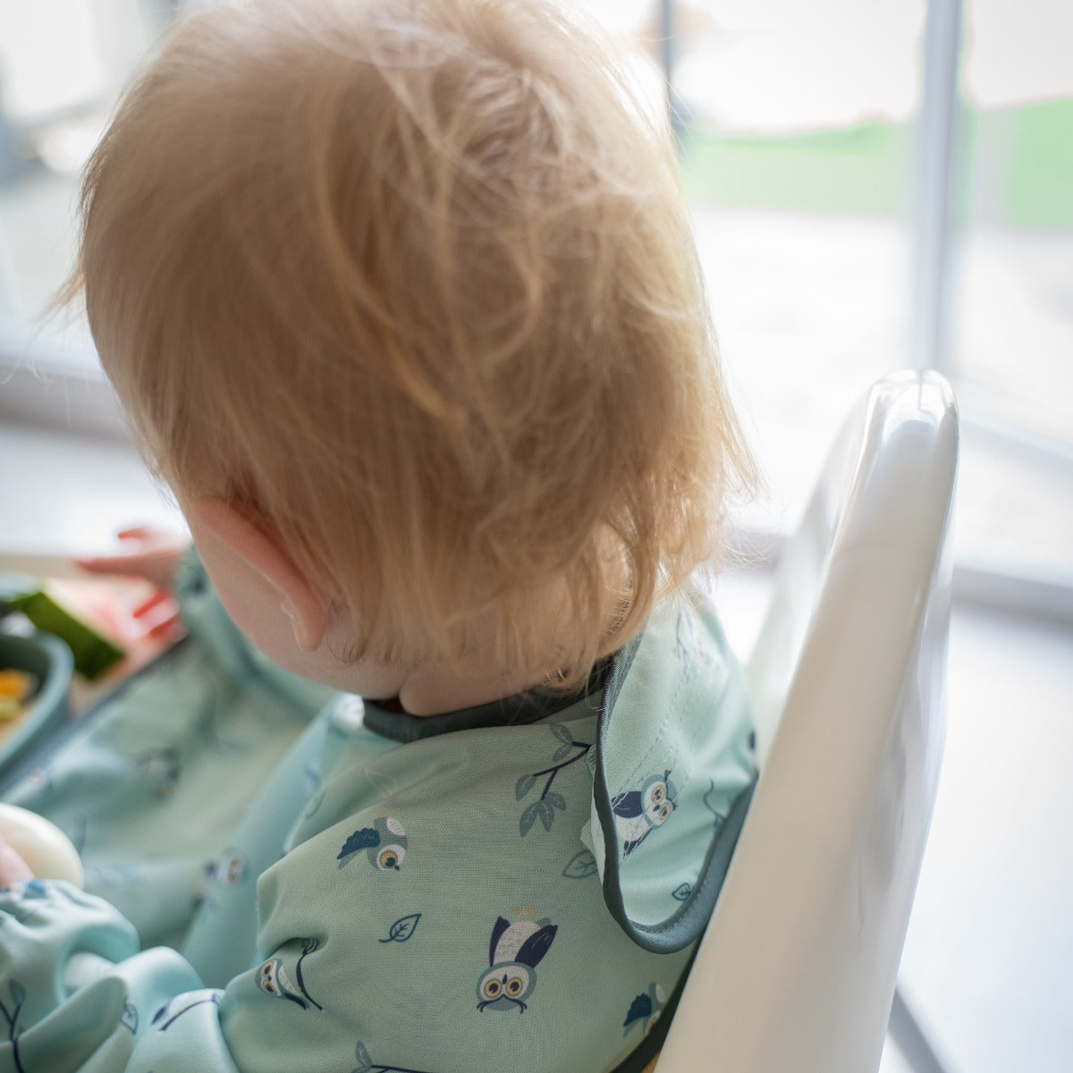Baby eating on a highchair wearing long sleeve bib with owl print rear view