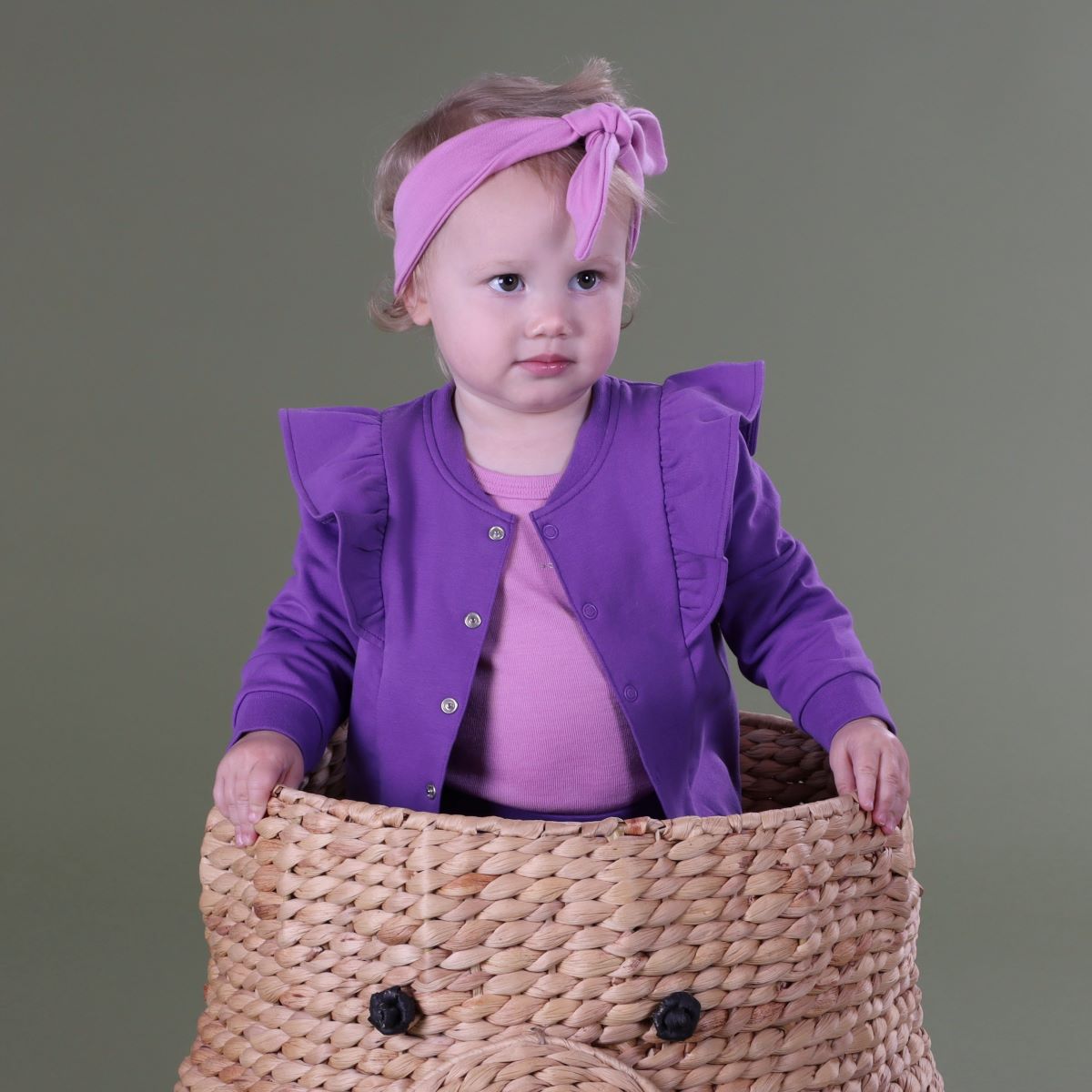 Baby girl standing inside a basket wearing Cotton Fleece Frill Jacket pairing it with pant, headband and merino top