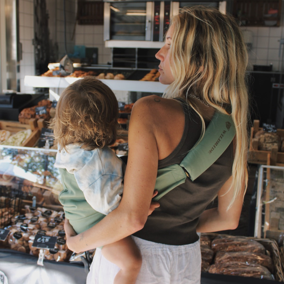 Mother at the store, holding toddler with Wildride Toddler Carrier in Fern colour

