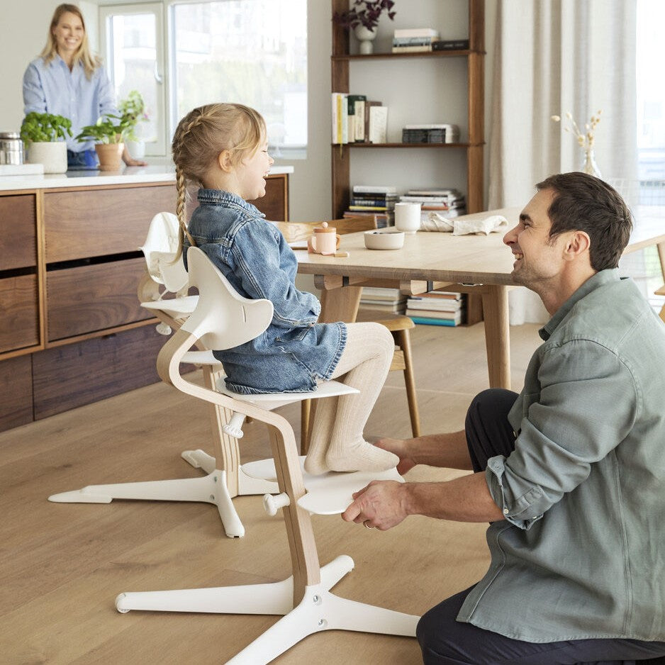 Baby girl sitting on a Nomi Natural white highchair from Stokke. and parents in dinning area