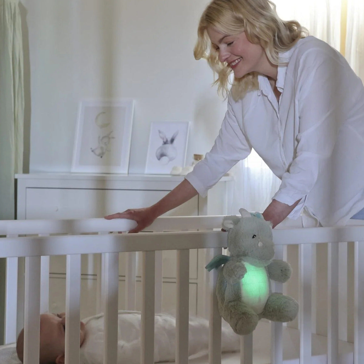Mother putting baby to bed with Cloud.b Lovelight Buddies - Drake the Dragon attached to cot