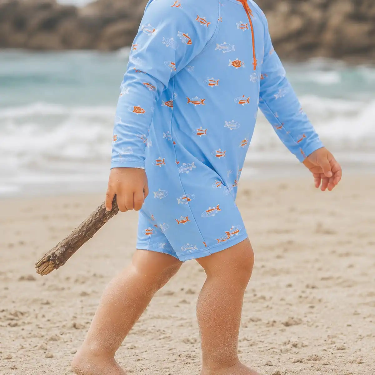 A child wearing a light blue long-sleeve swim cover-up with a small fish pattern walks on a sandy beach holding a piece of driftwood, with the ocean and rocks blurred in the background.