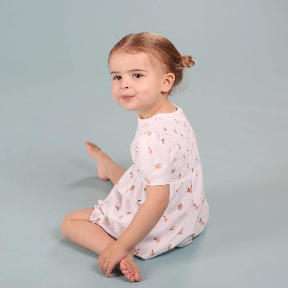 A toddler girl with light brown hair in a small bun sits on a light blue-gray studio background, wearing a pale pink short-sleeve romper with a peach pattern.