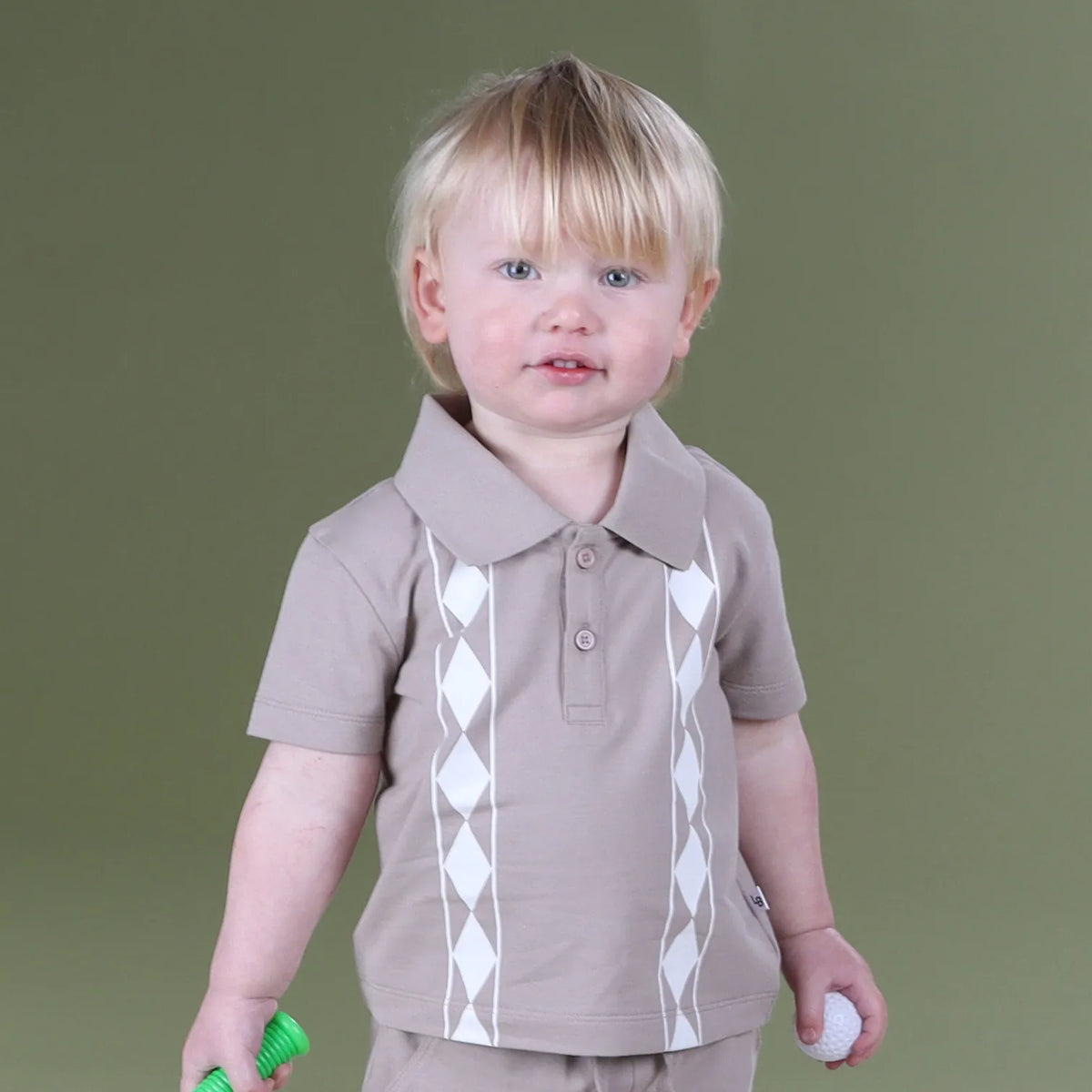 A toddler with blonde hair wearing a taupe short-sleeve polo shirt with white argyle diamond patterns on the front, standing against a solid olive green background.