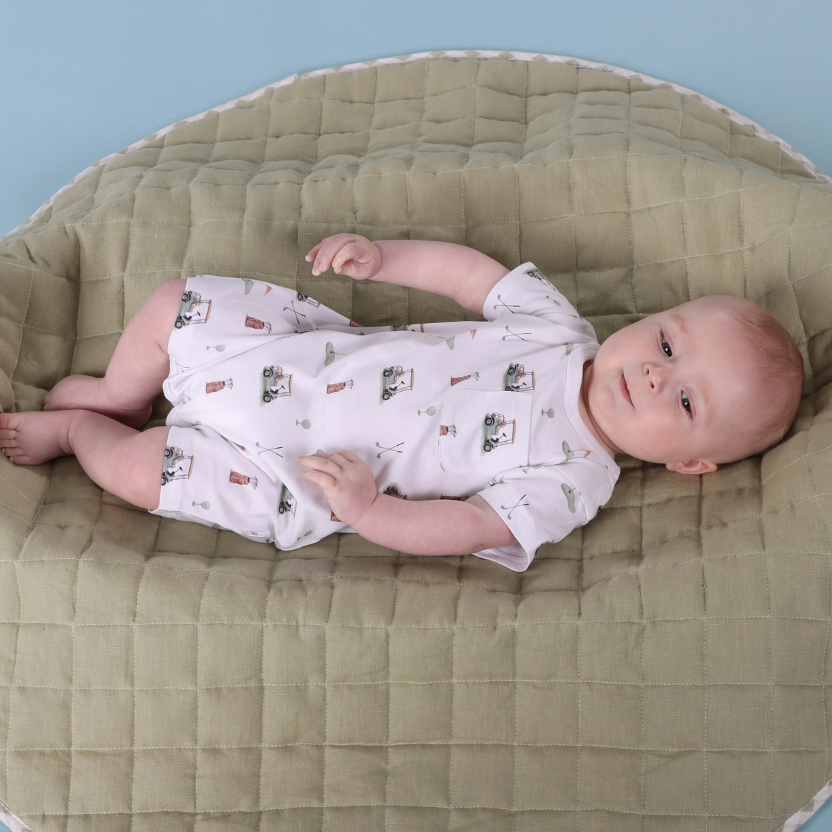 A baby lying on their back on a quilted olive green changing pad, wearing a white short-sleeve romper with a small vintage-style illustration pattern.