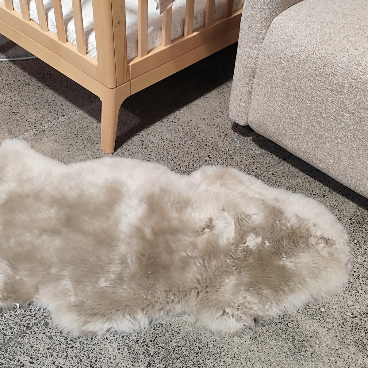 A fluffy sheepskin-style rug in a light beige and cream color lies on a speckled gray floor in a nursery or living room setting, with a wooden crib and a light-colored sofa visible in the background.