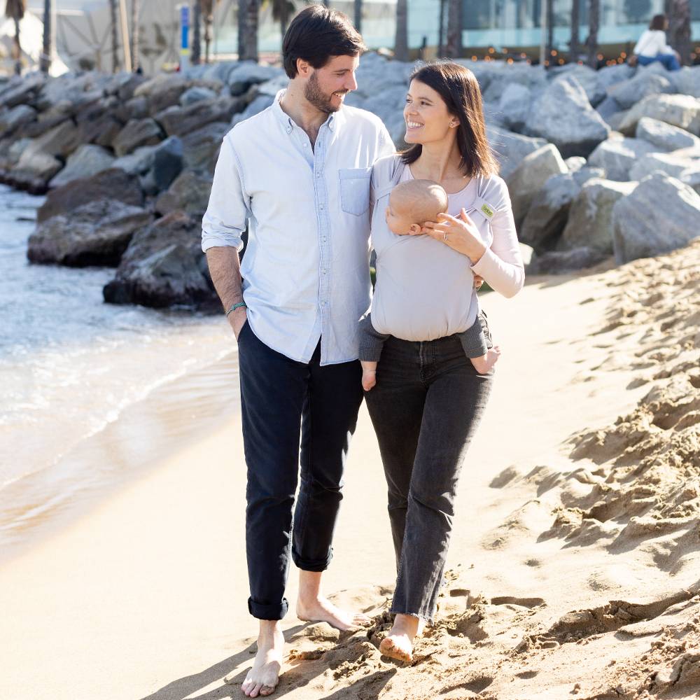 Couple walking along the beach with baby in Boba Bliss carrier in grey