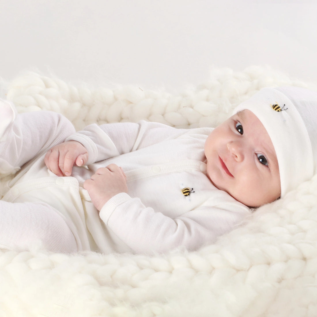 Baby in white outfit and hat lying on a white fluffy blanket