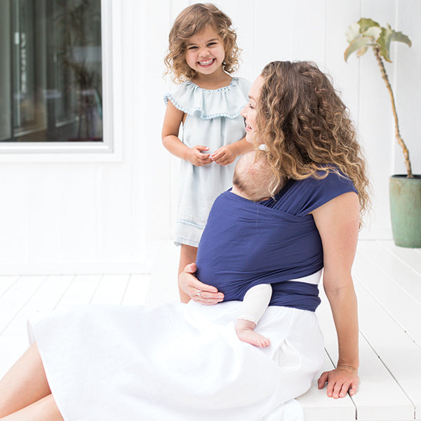 Mother sitting with a baby in a Ergobaby Indigo Aura Wrap and happy baby girl standing besides her.