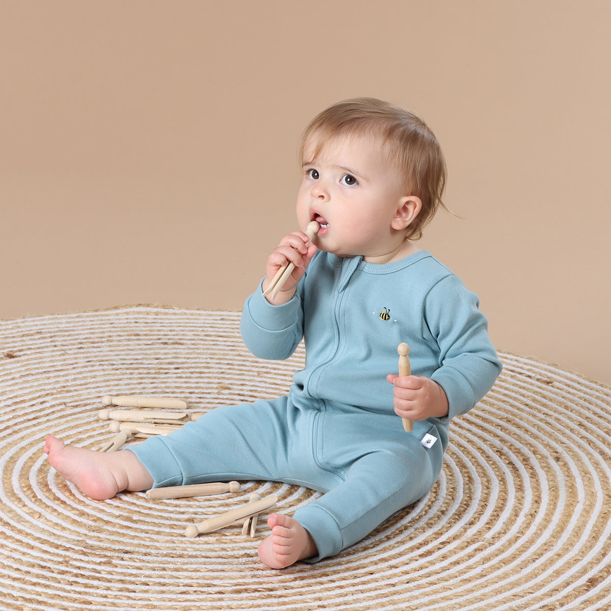 Baby holding pegs while wearing Cotton Zip long sleeve Babygrow with bee embroidery in ocean color