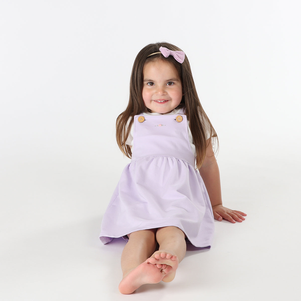 Playful baby girl wearing Cotton Pinafore dress for girls in lilac with rose embroidery and fliss and frankie bow headband