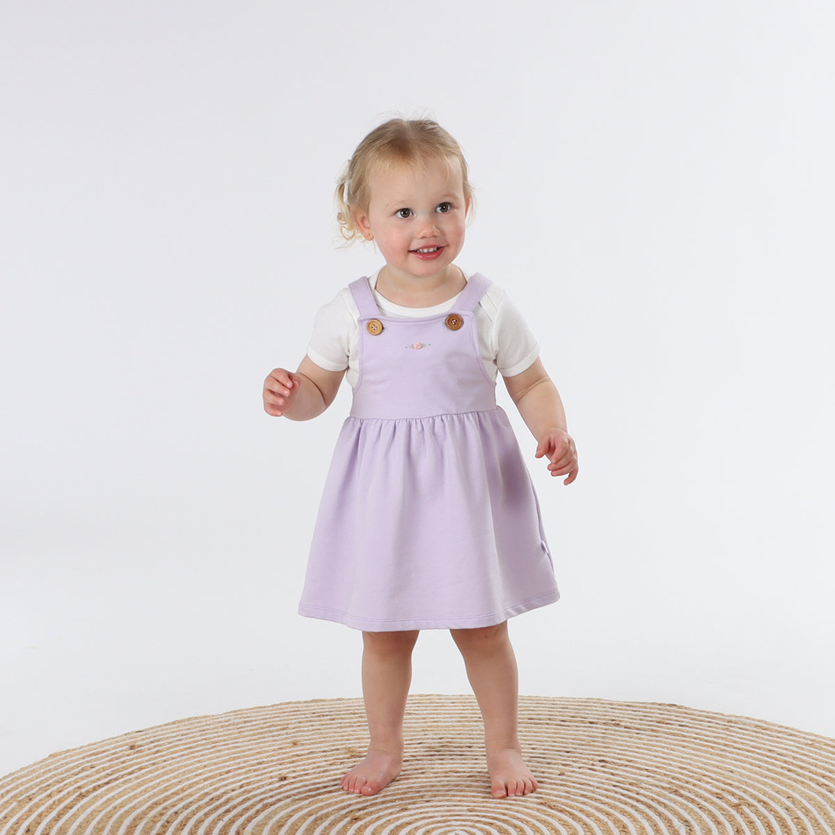 Playful baby girl wearing Cotton Pinafore dress for girls in lilac with rose embroidery and fliss and frankie bow headband