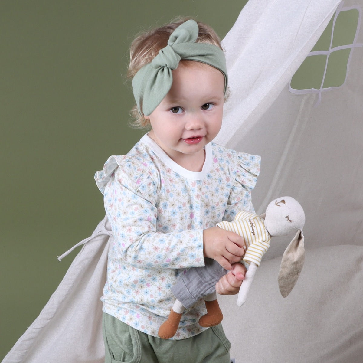 Baby girl wearing Cotton Frill Floral long sleeve top pairing it with merino headband and pant holding bunny in a tent