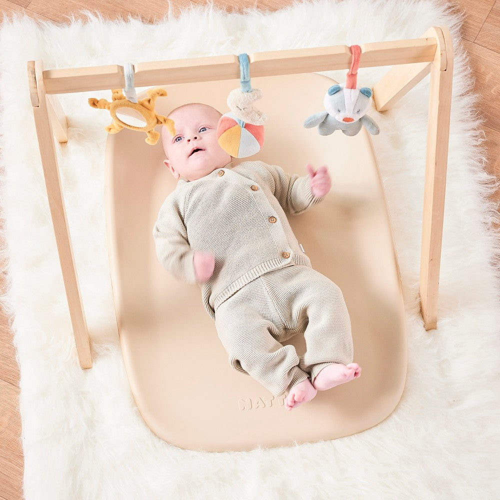 Baby palying with toy bar lying on Baby Change Mat from Nattou in beige