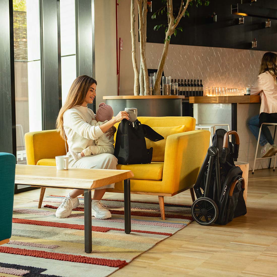 Mother in a cafe holding baby, sitting on a couch with folded Nuna Trvl Lx in Caviar