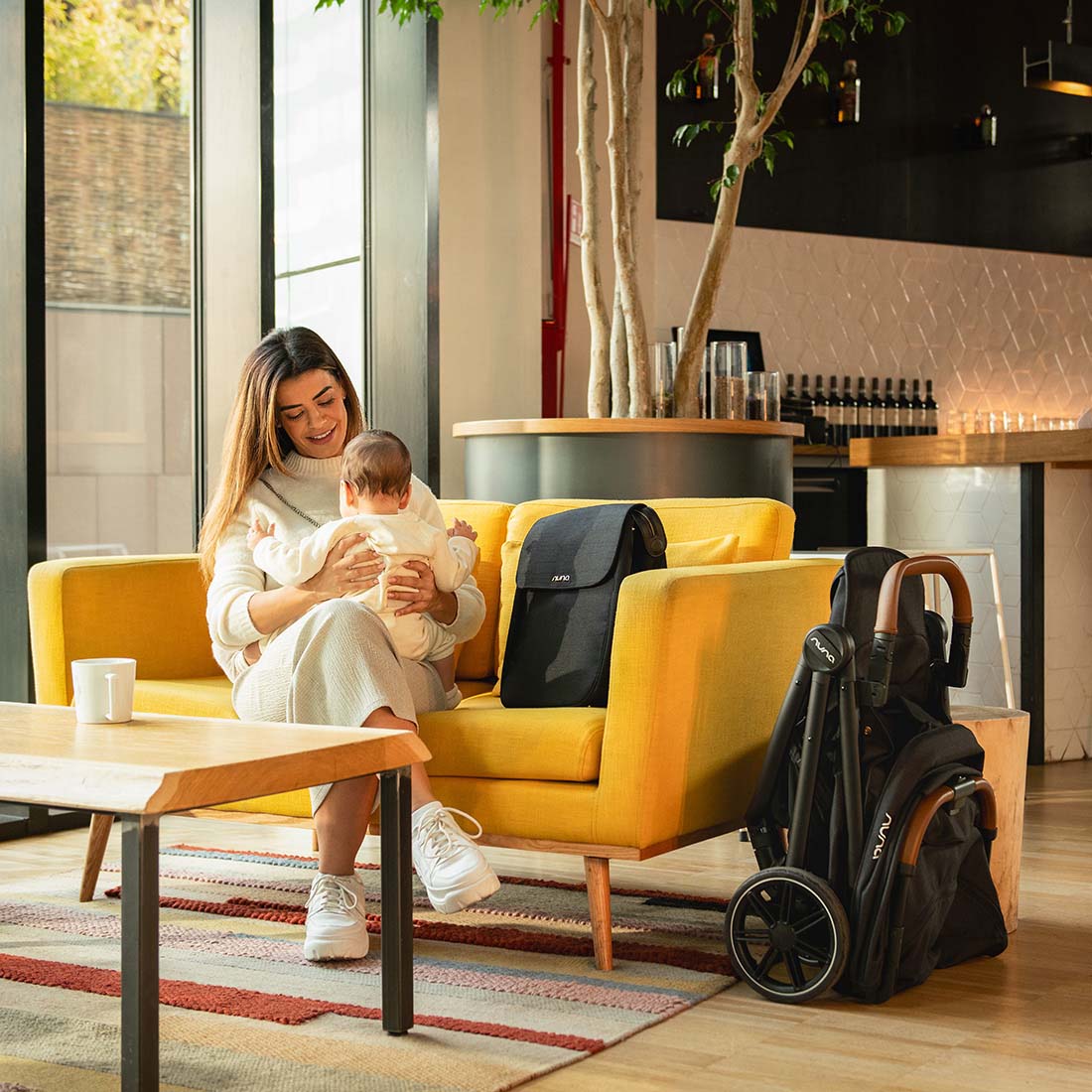 Mother in a cafe holding baby, sitting on a couch with folded Nuna Trvl Lx in Caviar
