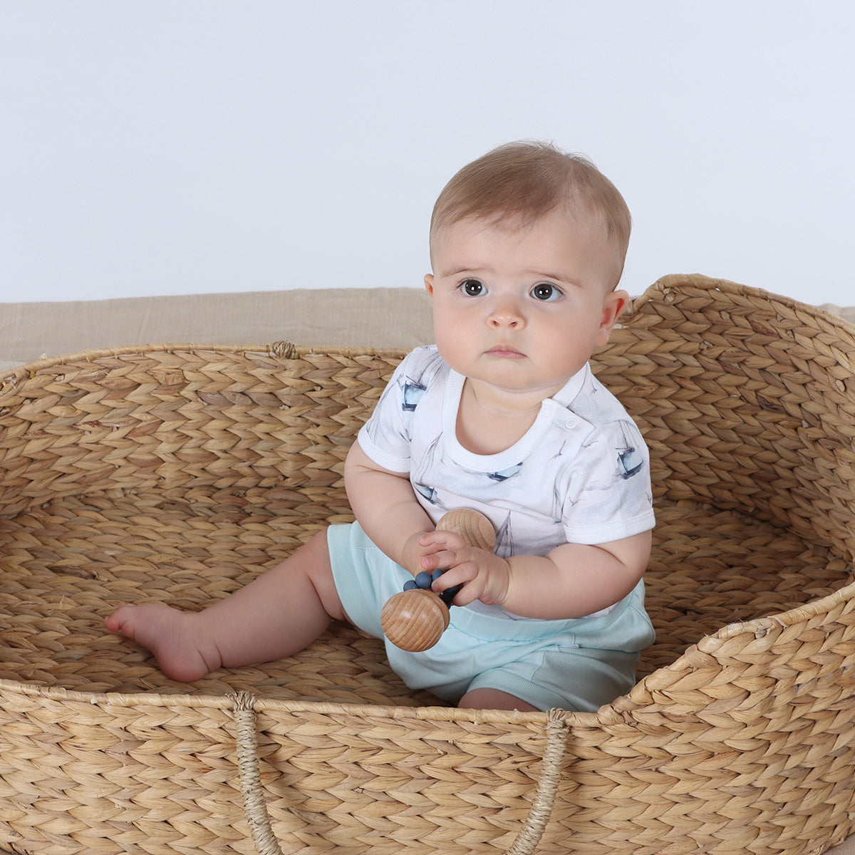 Baby playing with rattle wearing organic cotton bodysuit with boats print pairing it with blue shorts