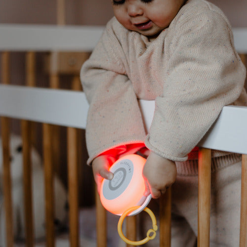 Baby in a cot holding Sleepy Tot portable white and pink noise machine with night light